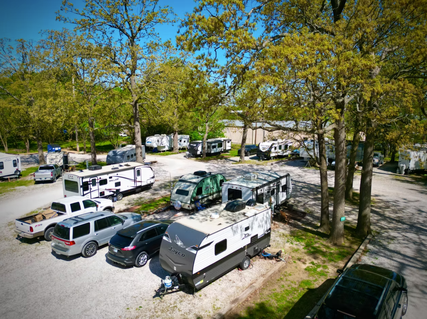 A gravel RV park nestled under lush green trees on a sunny day, filled with various campers, trucks, and parked cars.