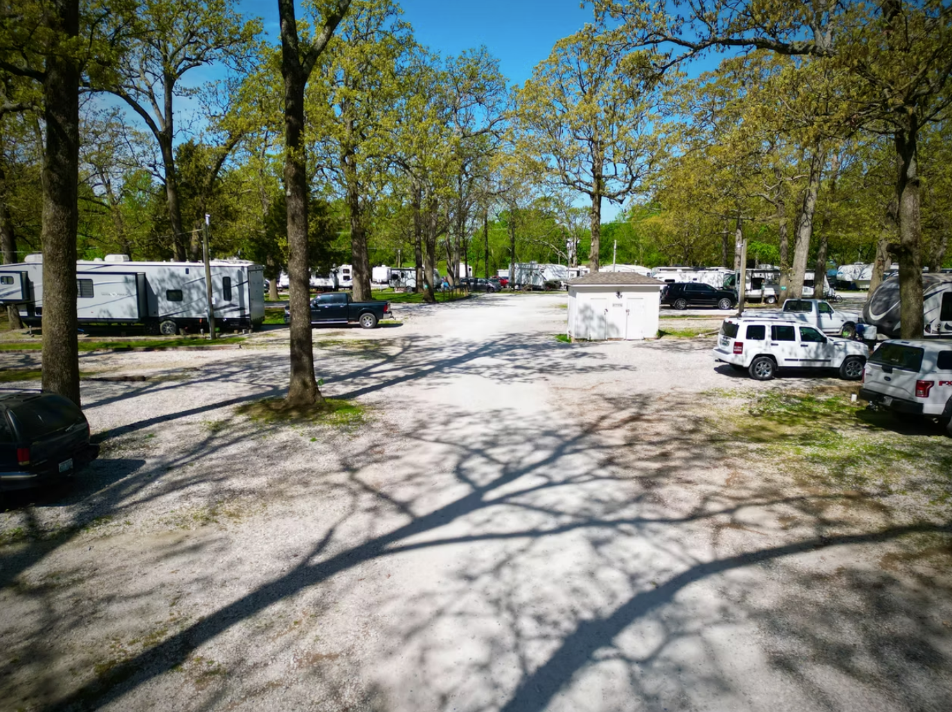 An outdoor RV park with gravel paths, mature trees casting long shadows, and various parked vehicles under a sunny sky.