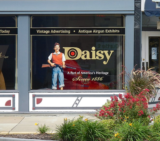 A storefront display featuring a Daisy logo, an airgun exhibit, and a cutout of a person holding a toy rifle.