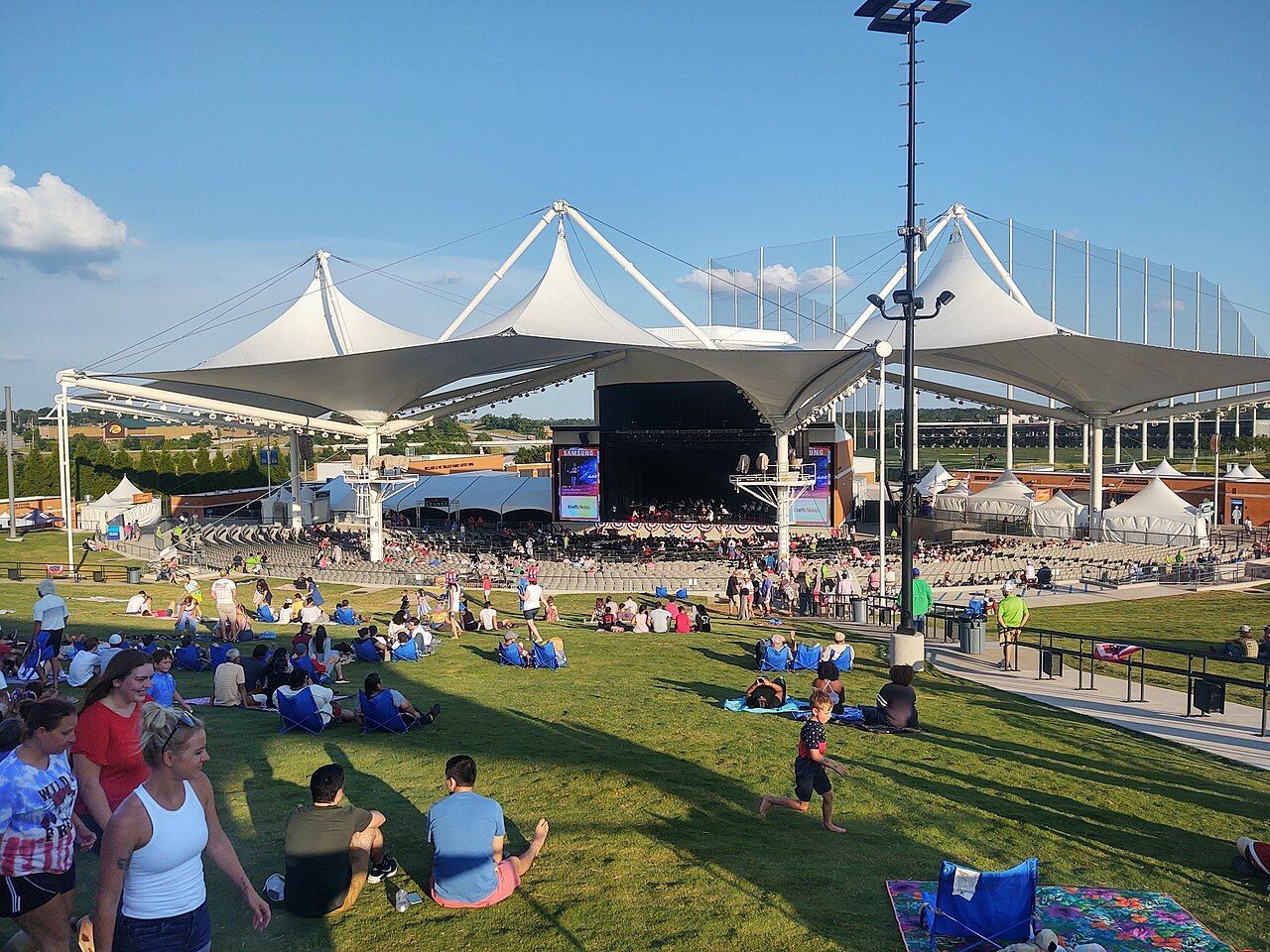 People sit on a grassy lawn at an outdoor amphitheater with white fabric tensile roofs under a clear blue sky.