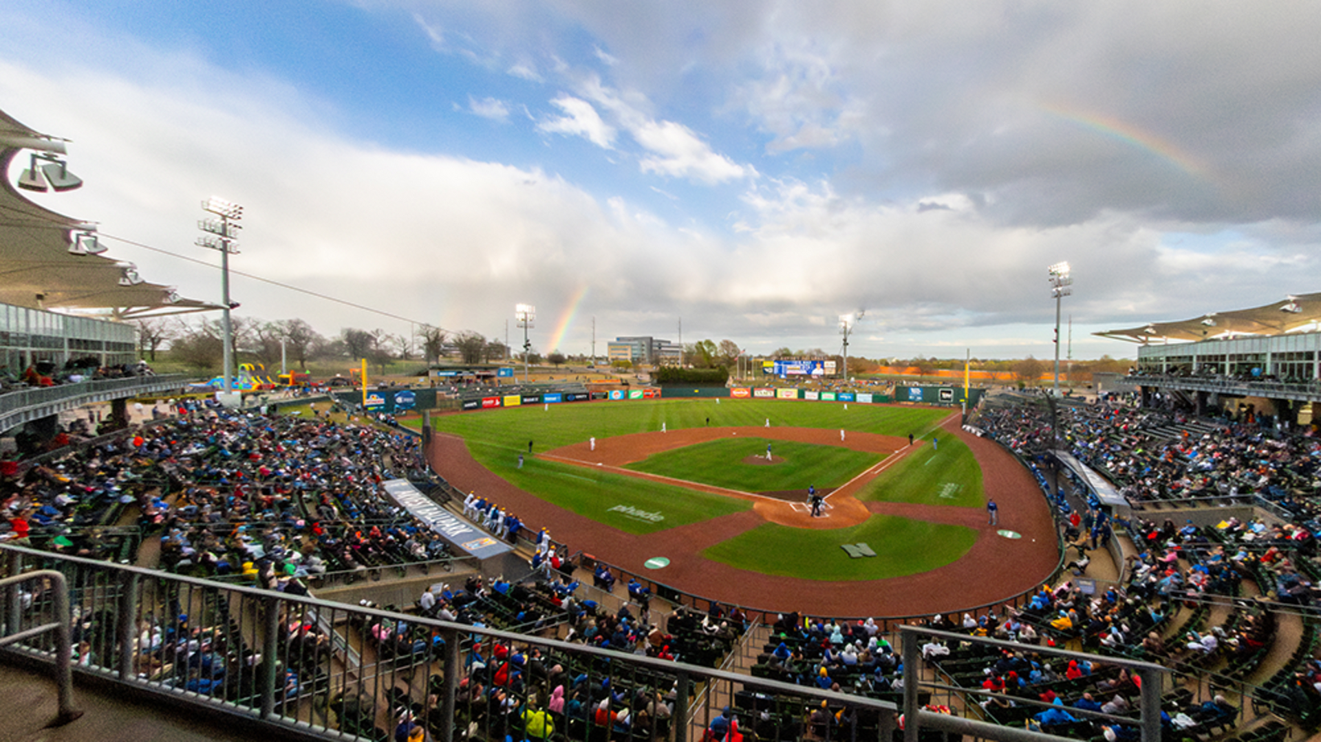 A high-angle view of a crowded baseball stadium under a cloudy sky featuring a faint rainbow over the outfield.