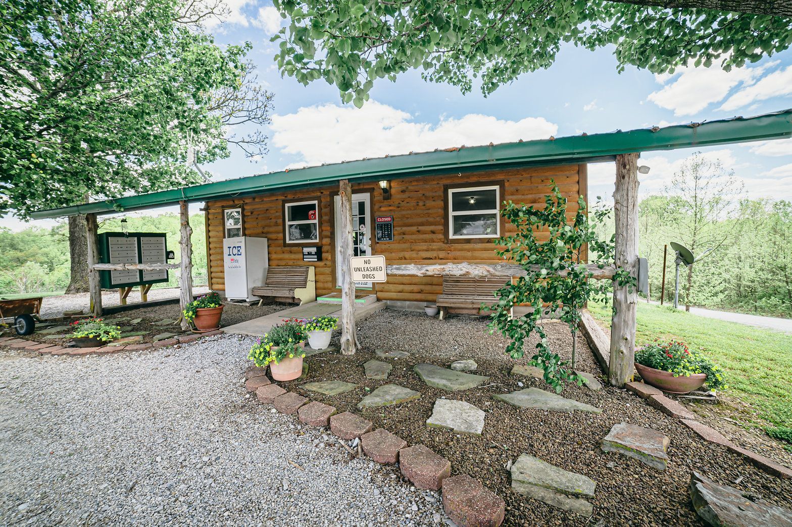 A small, wood-sided cabin with a wide, green-roofed porch, gravel driveway, and stone landscaping in a grassy, rural area.