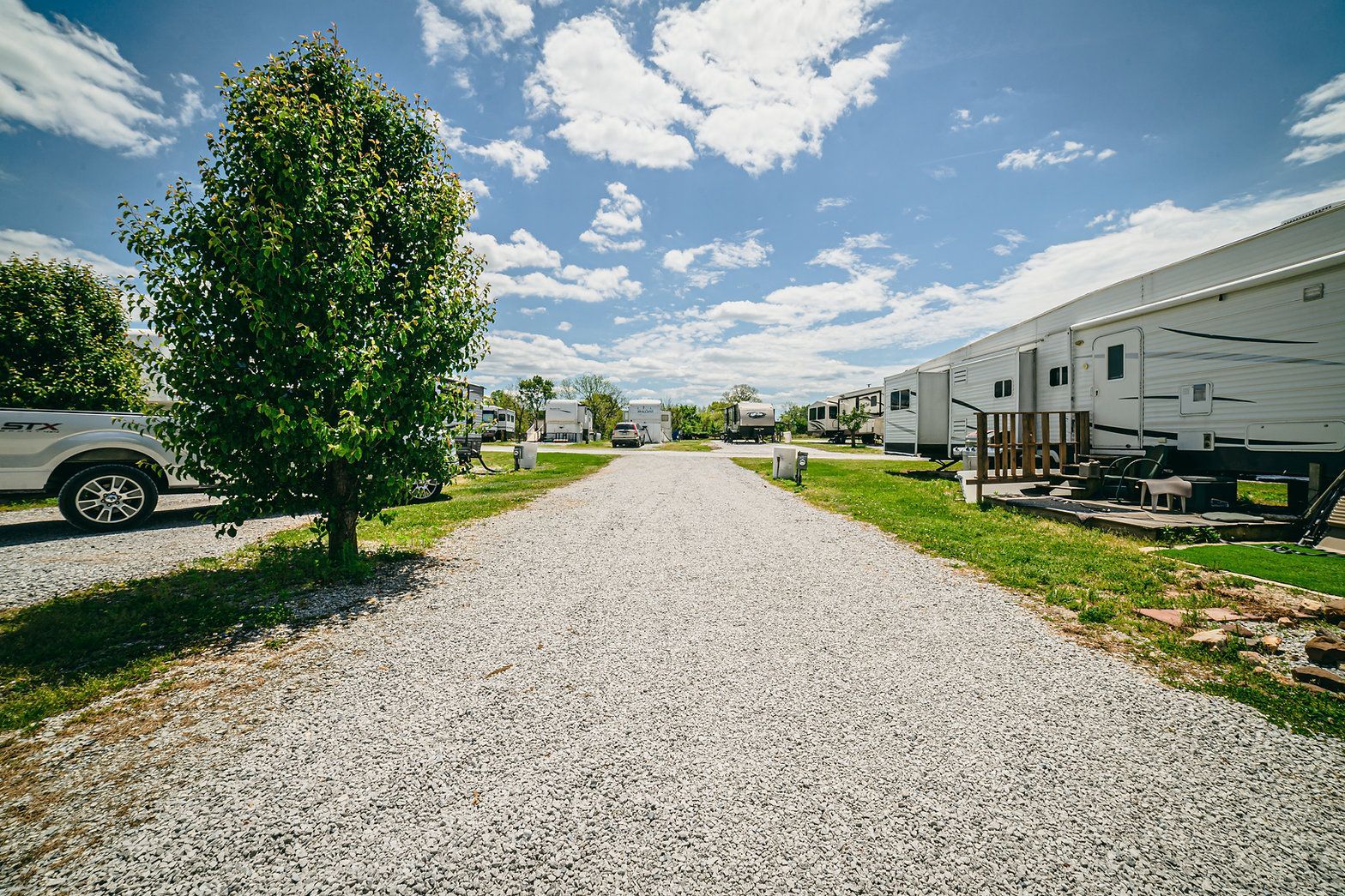 A gravel driveway leads through a sunny RV park lined with white campers and green trees under a blue, cloudy sky.