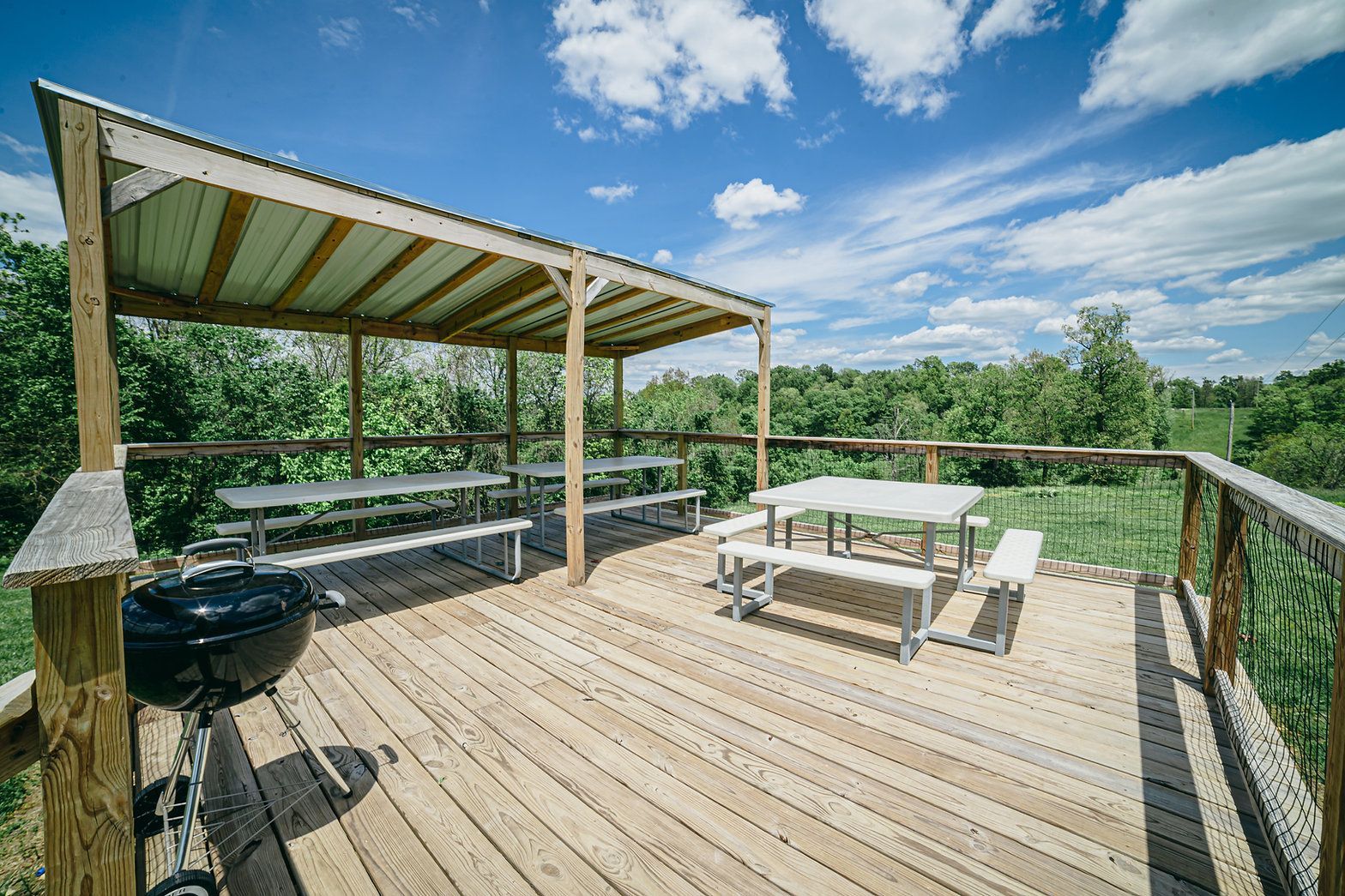 A wooden deck with two picnic tables, one covered by a shed roof, and a charcoal grill on a sunny day.