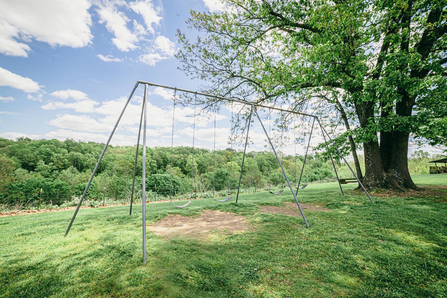 An old, empty metal swing set stands on a grassy hill near a large tree under a blue sky with scattered clouds.