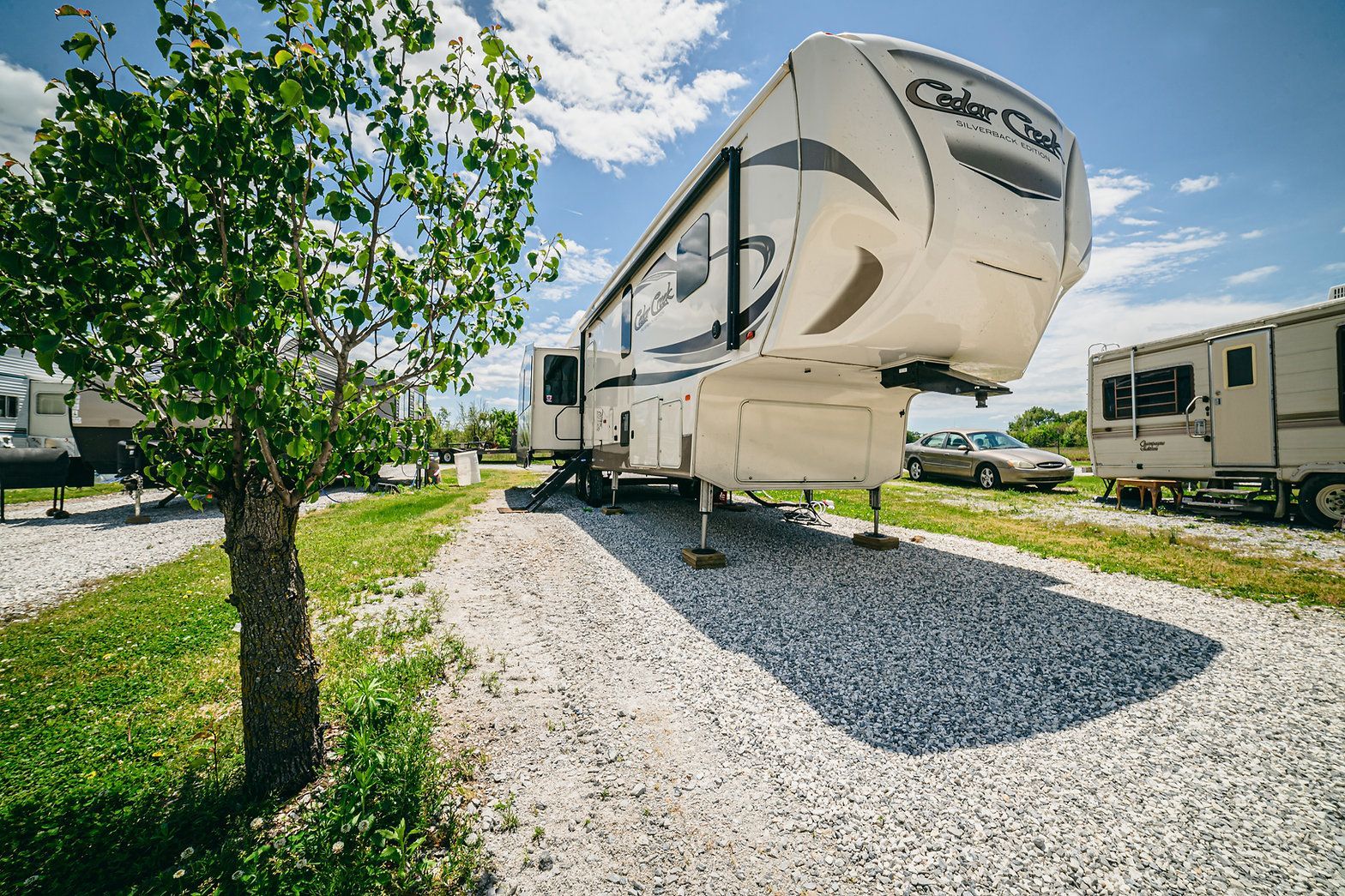 A white Cedar Creek fifth-wheel RV parked on a gravel lot next to a small tree under a sunny, blue sky.