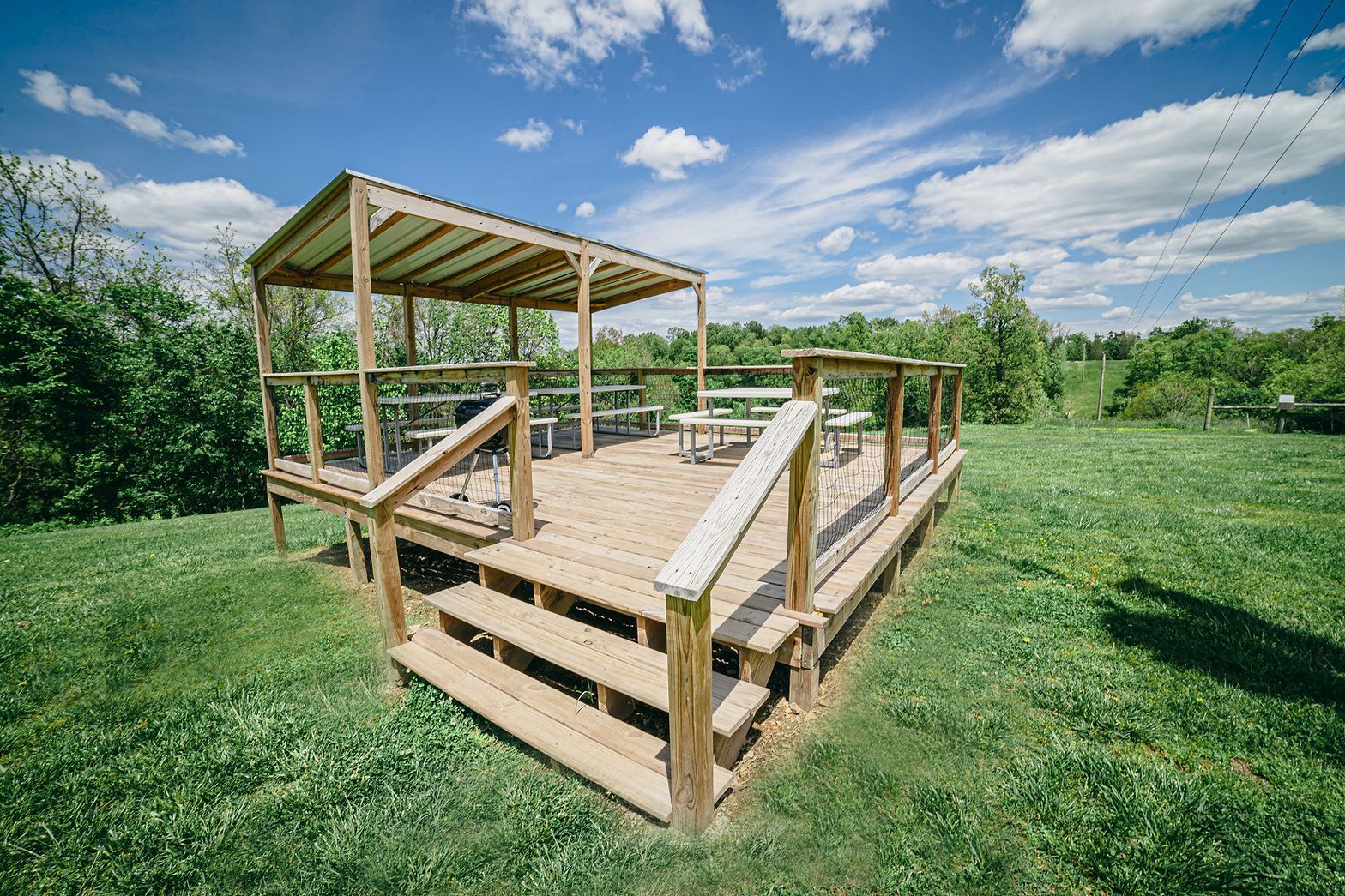 A wooden deck with a partially covered roof and seating area, situated in a grassy field under a bright blue sky.