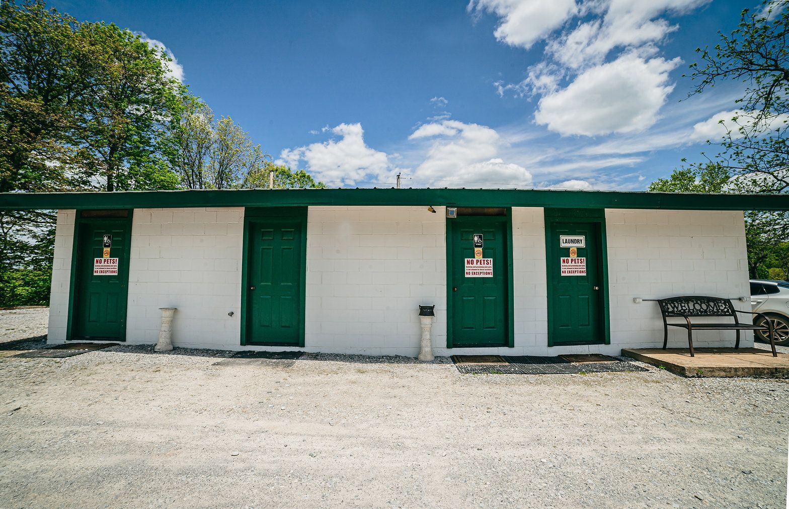 A white building with a green roof and four green doors, each featuring a sign, sitting on a gravel lot under a blue sky.