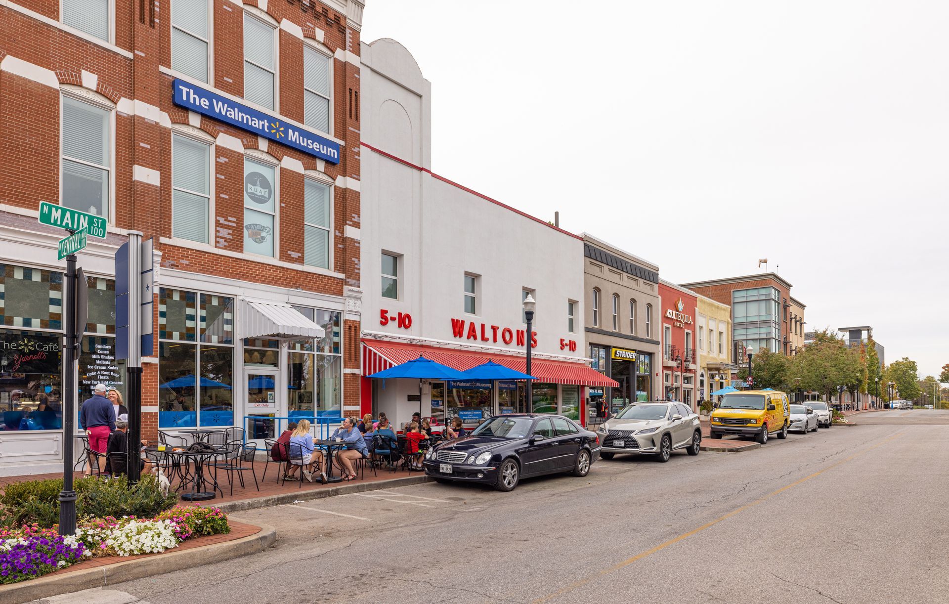 A street scene featuring red brick and white storefronts, including a Walton’s 5-10, with sidewalk seating and parked cars.