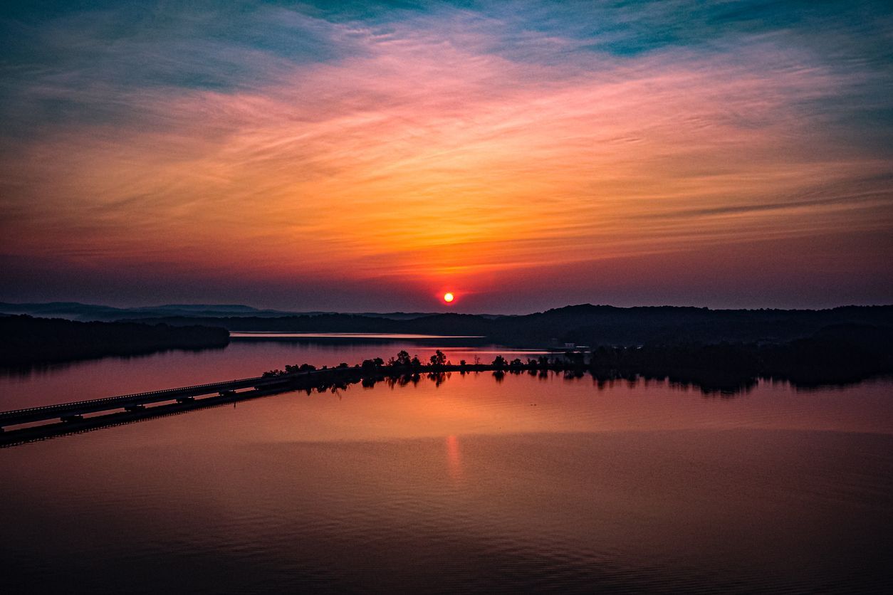 A sunset glows orange and pink over a dark, calm lake, reflecting the light with a bridge crossing the water.
