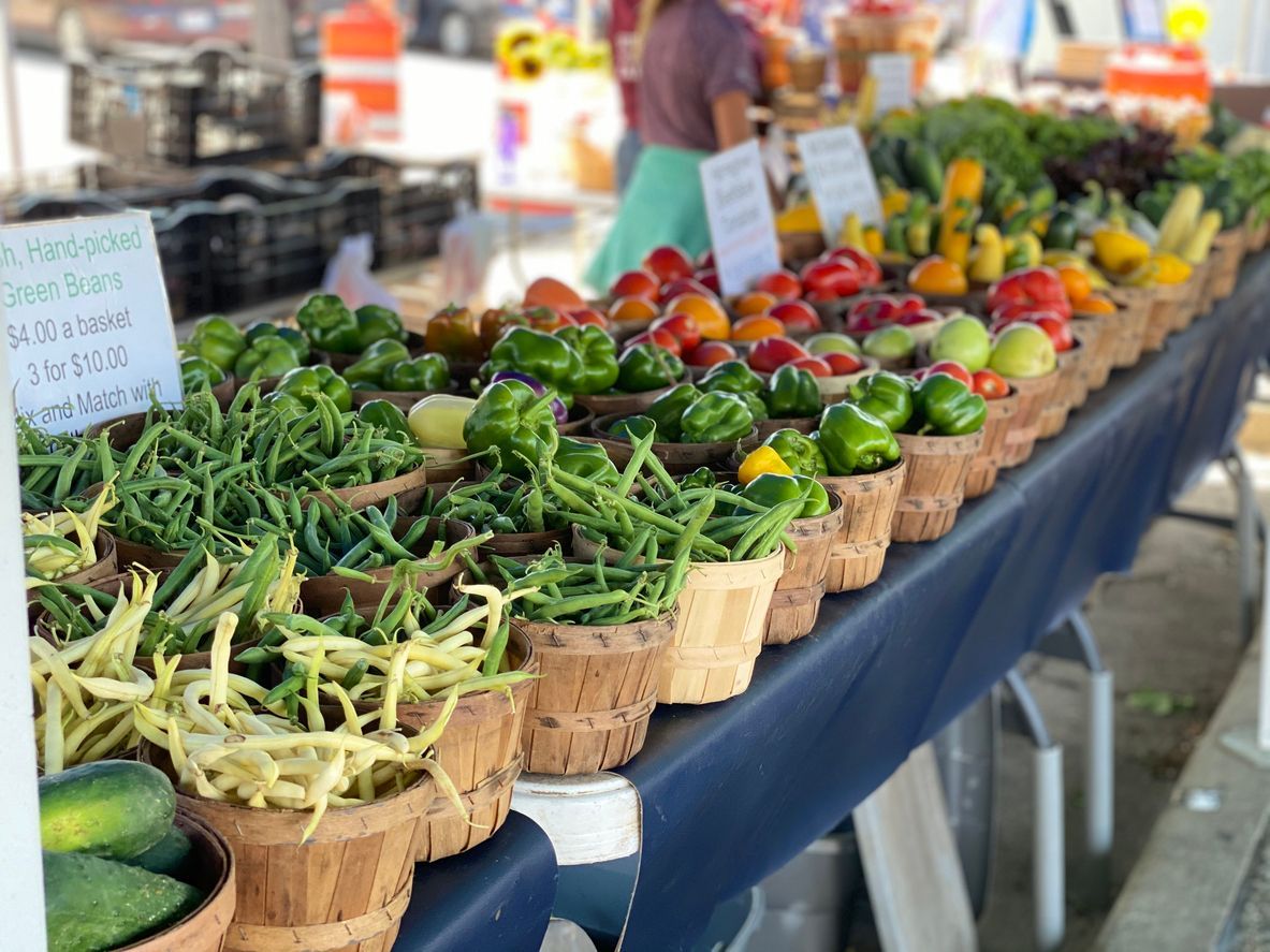 A farmers market stall displays rows of baskets filled with fresh green beans, peppers, and tomatoes on a blue table.