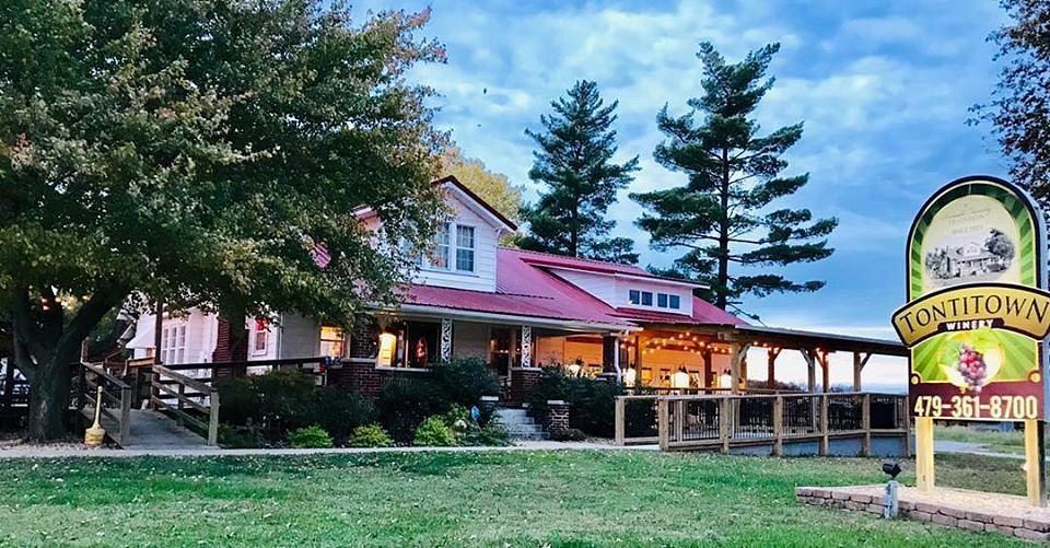 Honeytown winery building with a red roof, front porch, and tall trees, alongside a business sign on a sunny day.