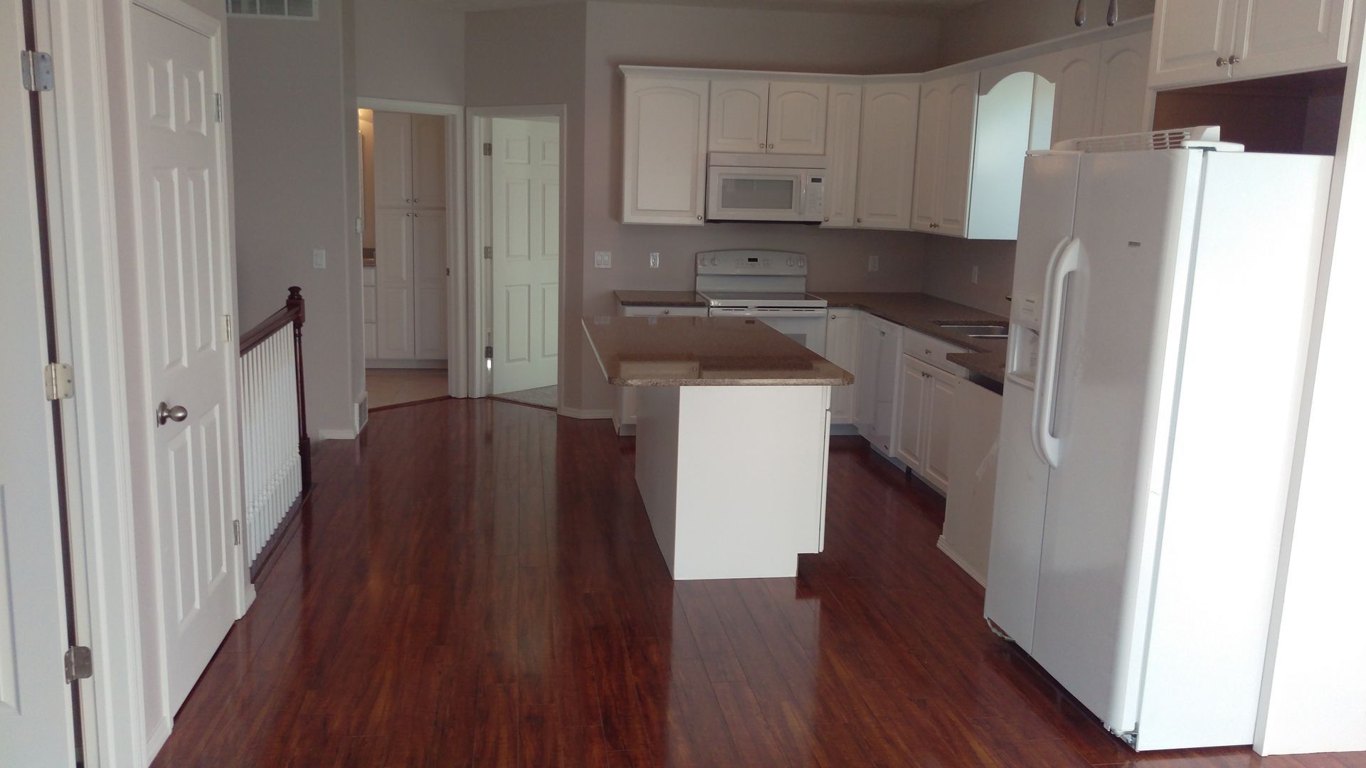 An empty kitchen with hardwood floors and white cabinets and a refrigerator.