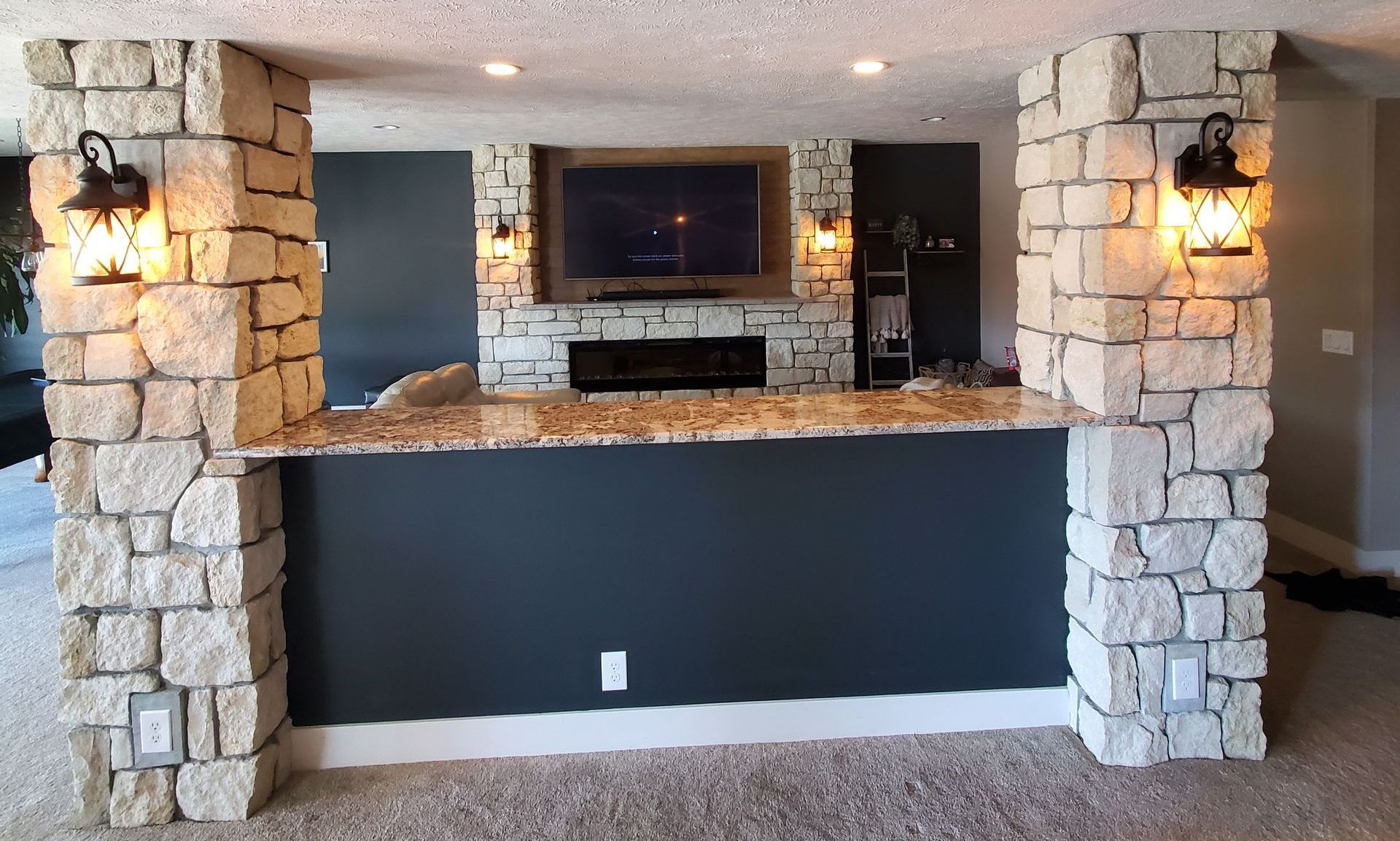 Stone-faced bar with a built-in TV and fireplace. The backdrop is navy blue, with sconces and a carpeted floor.