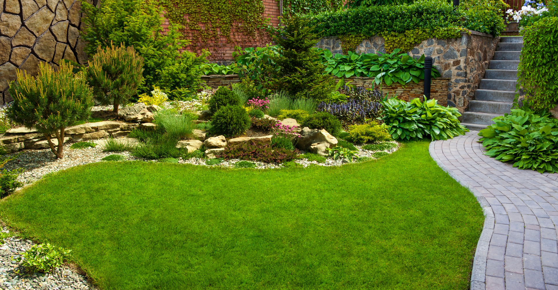 A lush green garden with a brick walkway leading to a stone wall and stairs.