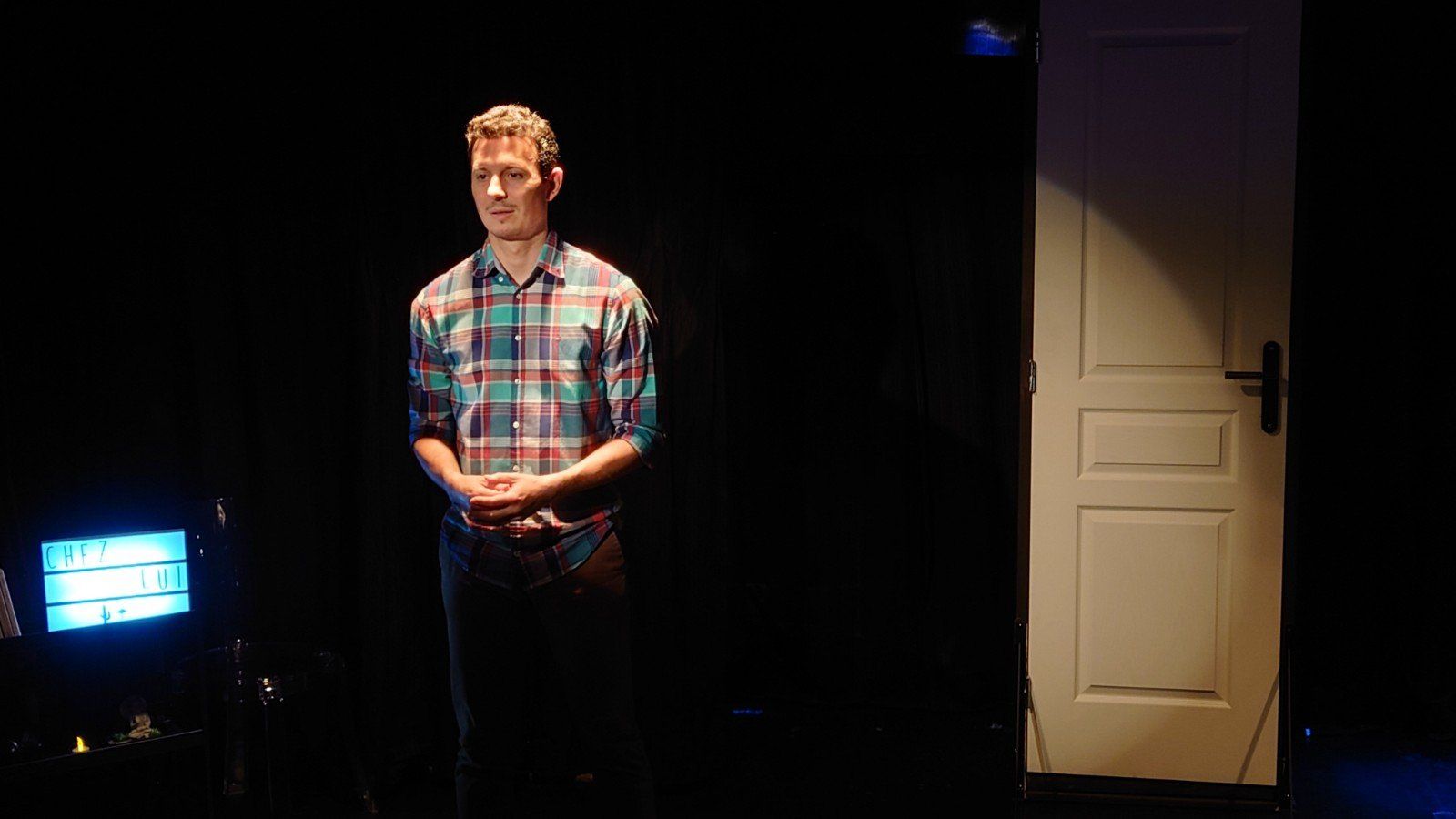 un homme vêtu d'une chemise à carreaux se tient devant une porte blanche dans une pièce sombre.