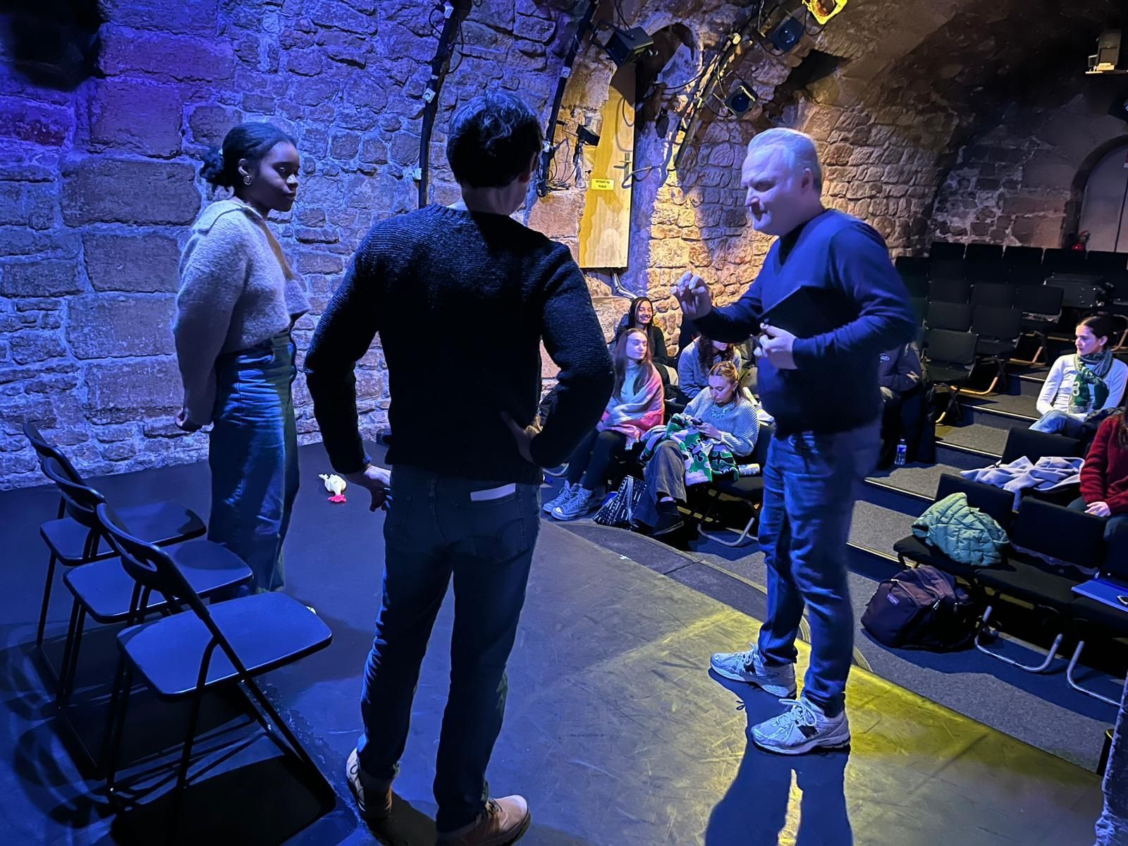 Trois personnes dans une pièce aux murs de pierre ; l’une fait un geste, les deux autres observent. Public en arrière-plan.