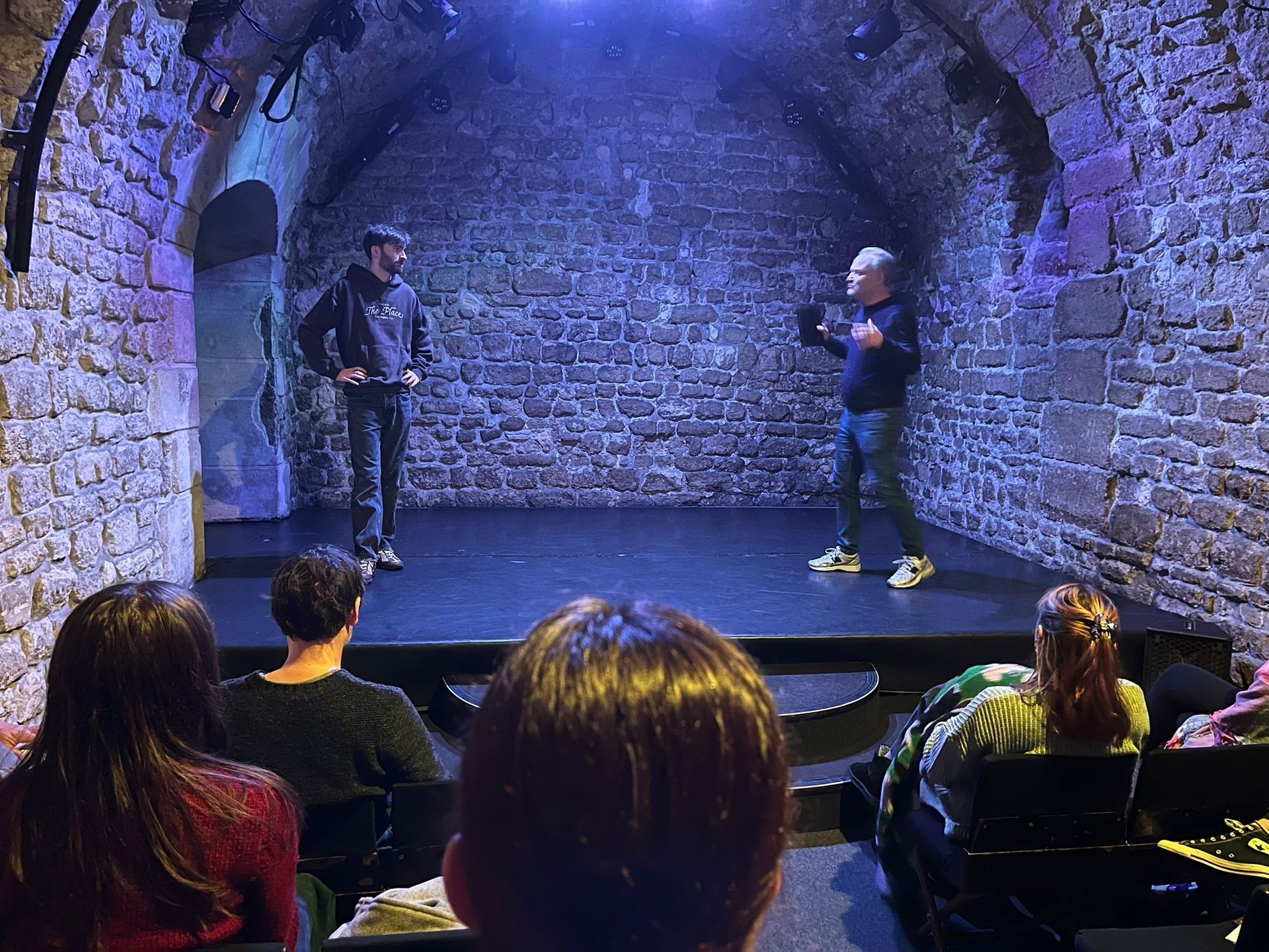 Deux personnes sur une petite scène, devant un public, dans une salle aux murs de pierre.