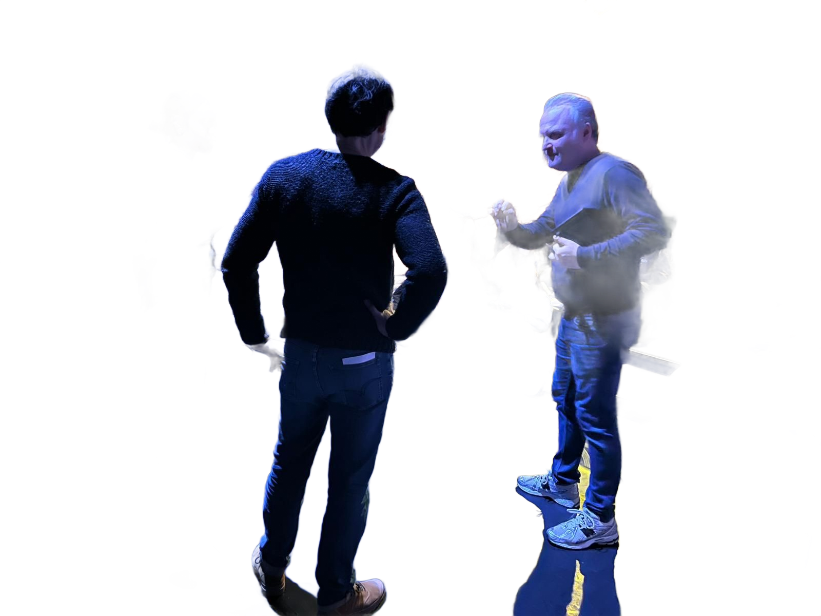 Dans une salle de spectacle : un homme fait un geste, deux autres sont debout. Chaises et public en arrière-plan.