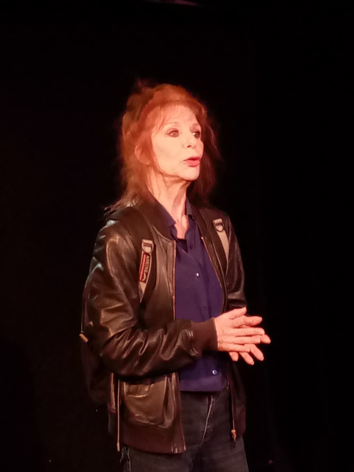Une femme portant une veste en cuir et une chemise bleue se tient debout sur une scène.