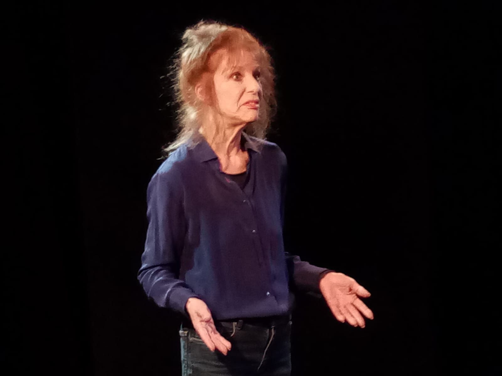 Une femme en chemise bleue se tient debout sur une scène avec les bras tendus.