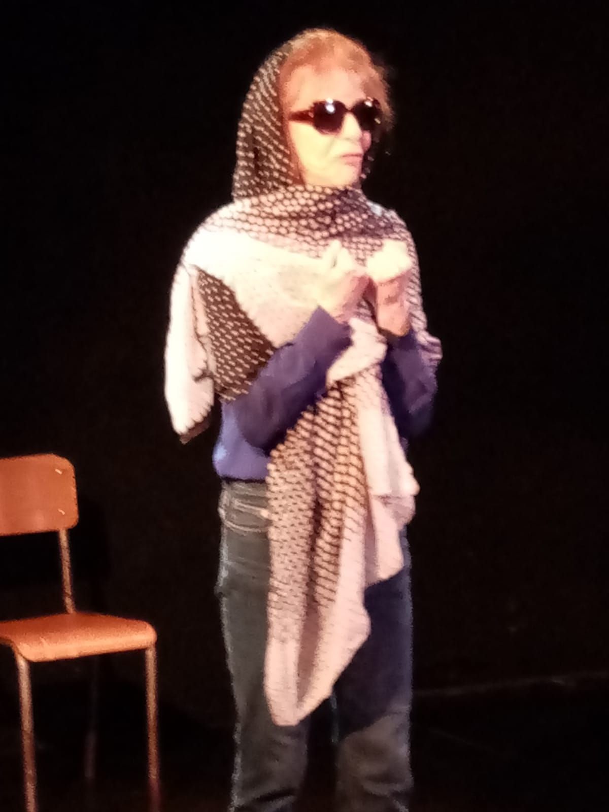 Une femme portant des lunettes de soleil et un foulard se tient devant une chaise