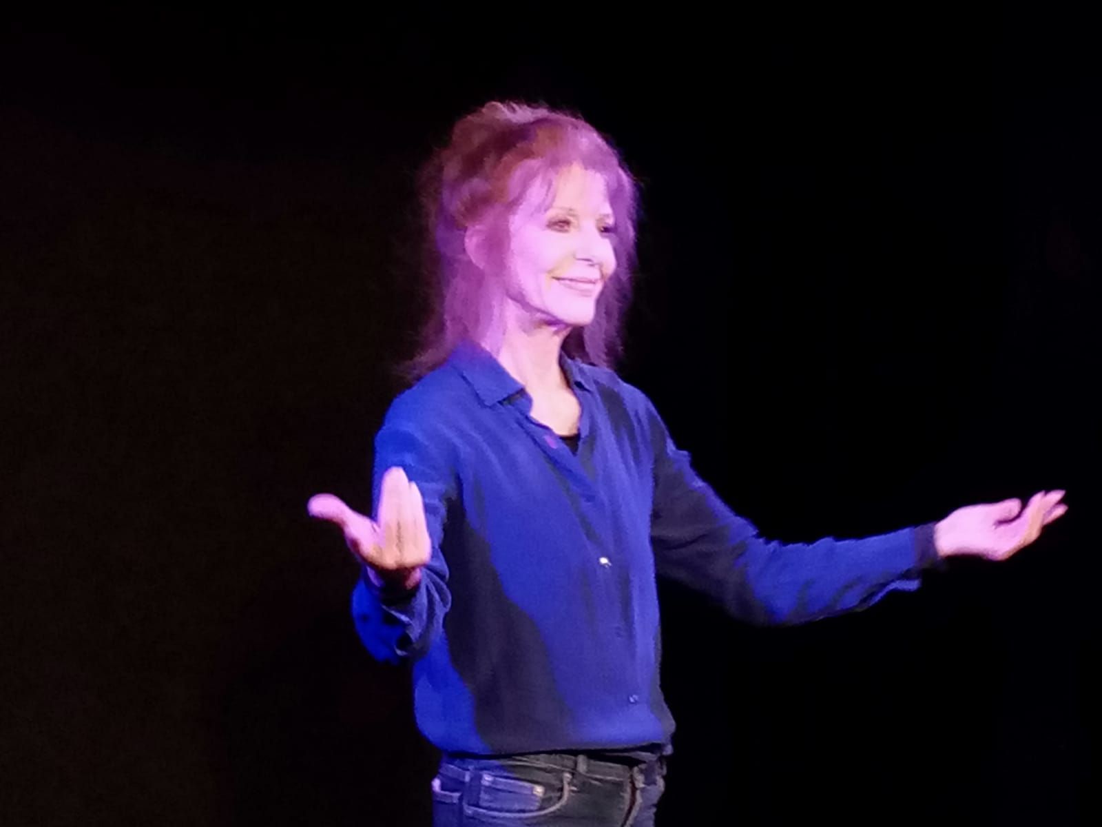 Une femme en chemise bleue se tient debout sur une scène avec les bras tendus.