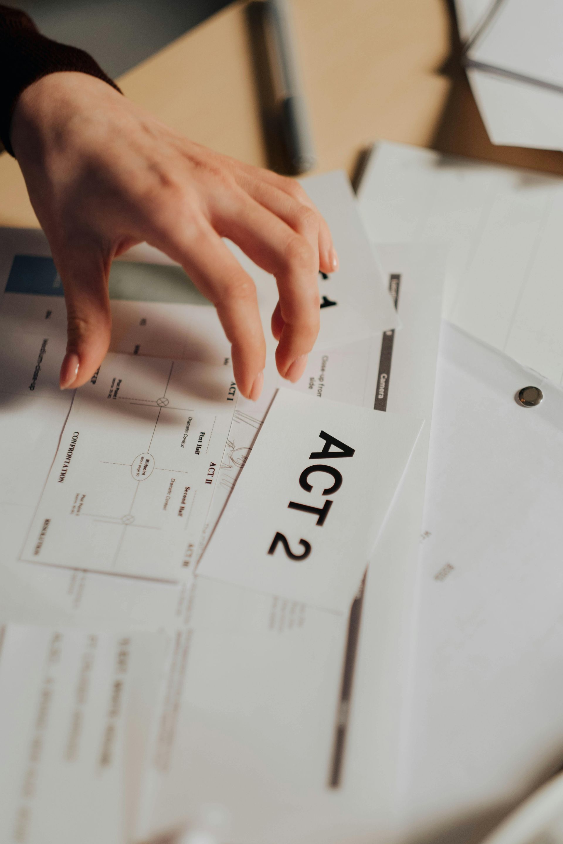 Käsi kurottaa kohti "Näytös 2":n käsikirjoituksen sivua, joka on papereilla peitetyllä pöydällä.