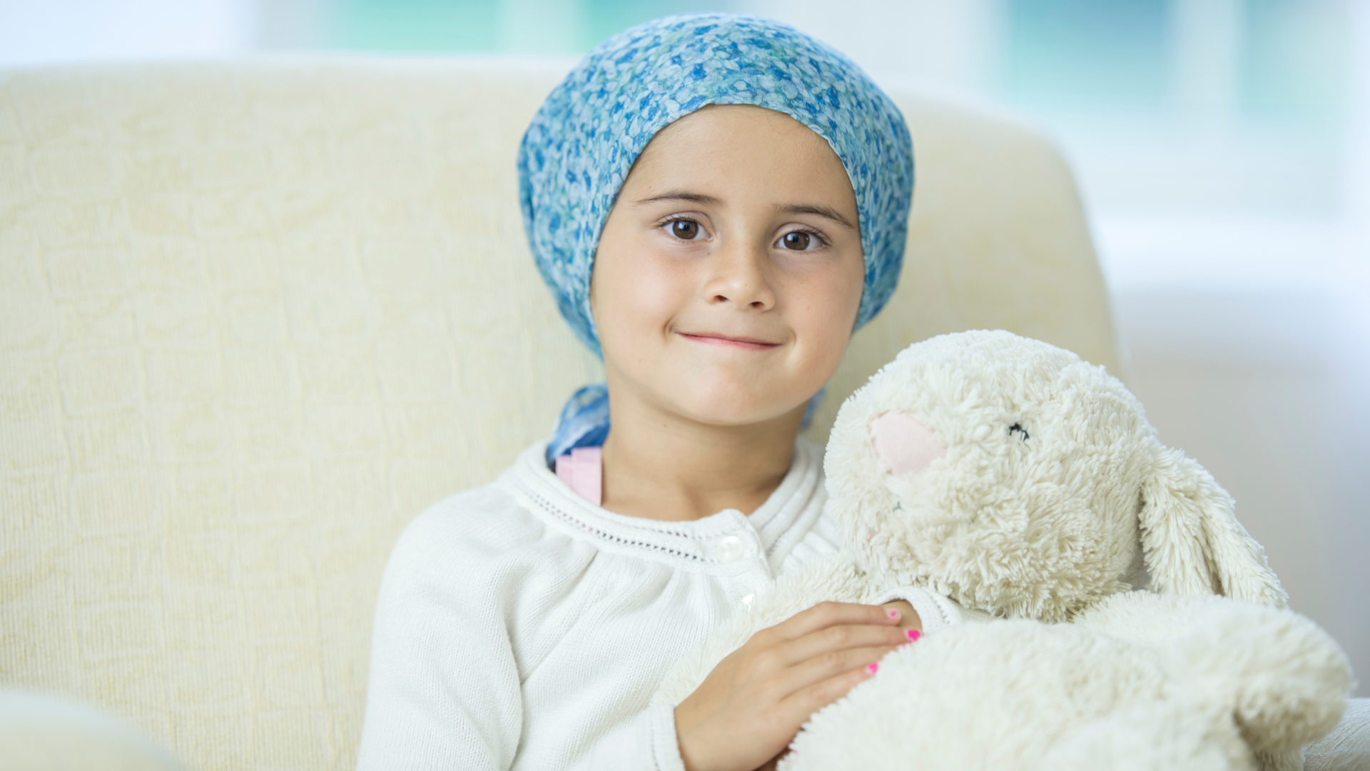 une petite fille atteinte d'une leucémie assise sur un fauteuil avec une peluche et un foulard