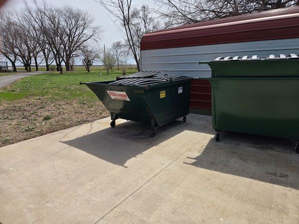 A large blue dumpster filled with wooden debris, branches, and discarded items outdoors under trees.