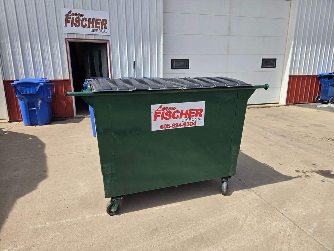 A person in dark clothing maneuvers a large green wheeled bin on a paved path between buildings.