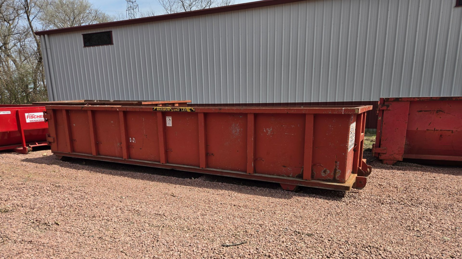 A large, empty blue dumpster sits on a concrete driveway in front of a house, surrounded by a lawn and trees.
