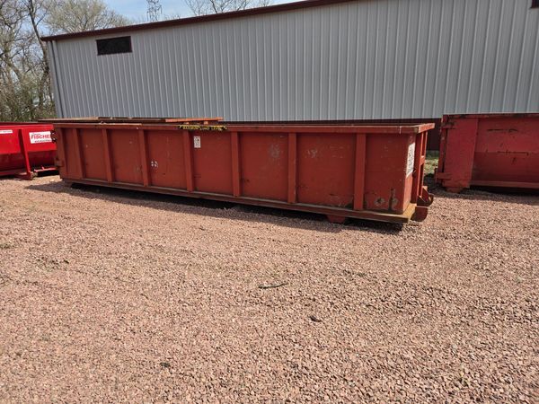 A large, empty bright red metal dumpster sits on an asphalt road in front of lush green trees.