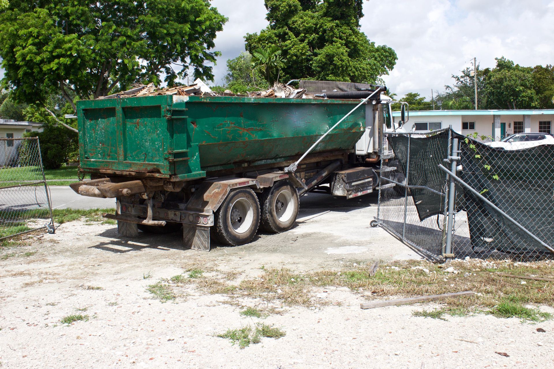 A large green dumpster truck parked on a gravel lot near a chain-link fence, viewed from the rear.