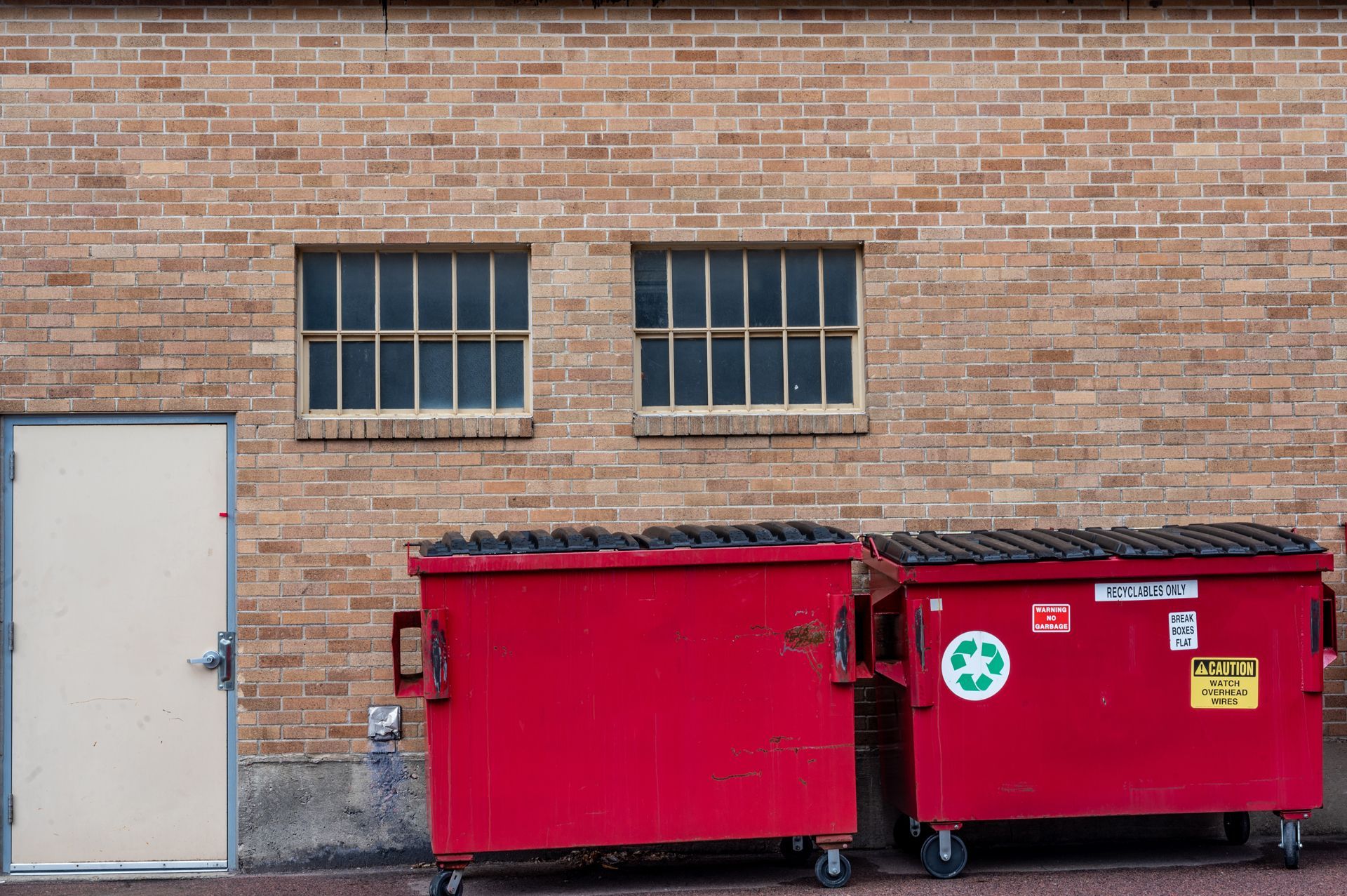 Two red dumpsters sit in front of a brick building with two windows and a white door.