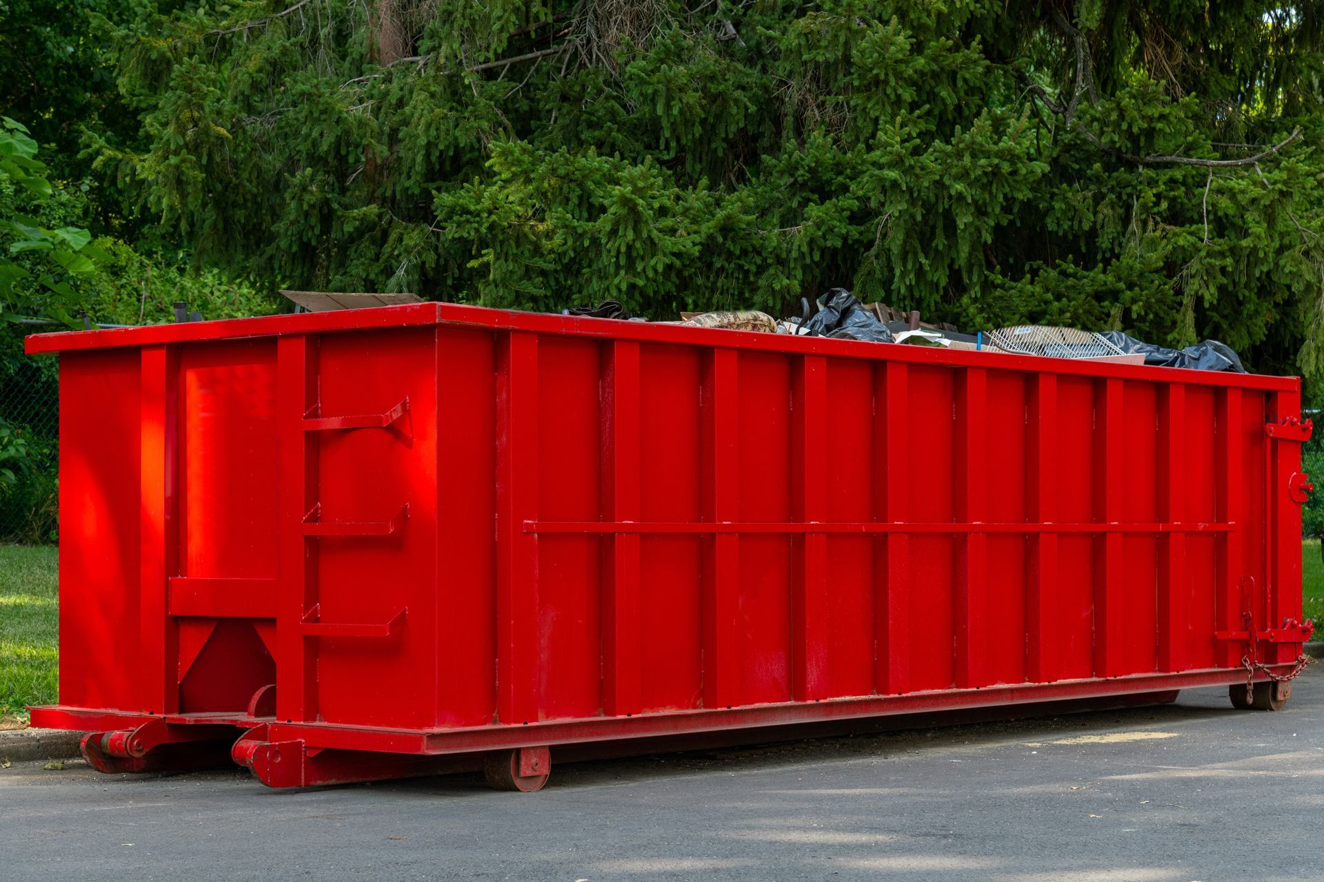 A large, empty bright red metal dumpster sits on an asphalt road in front of lush green trees.