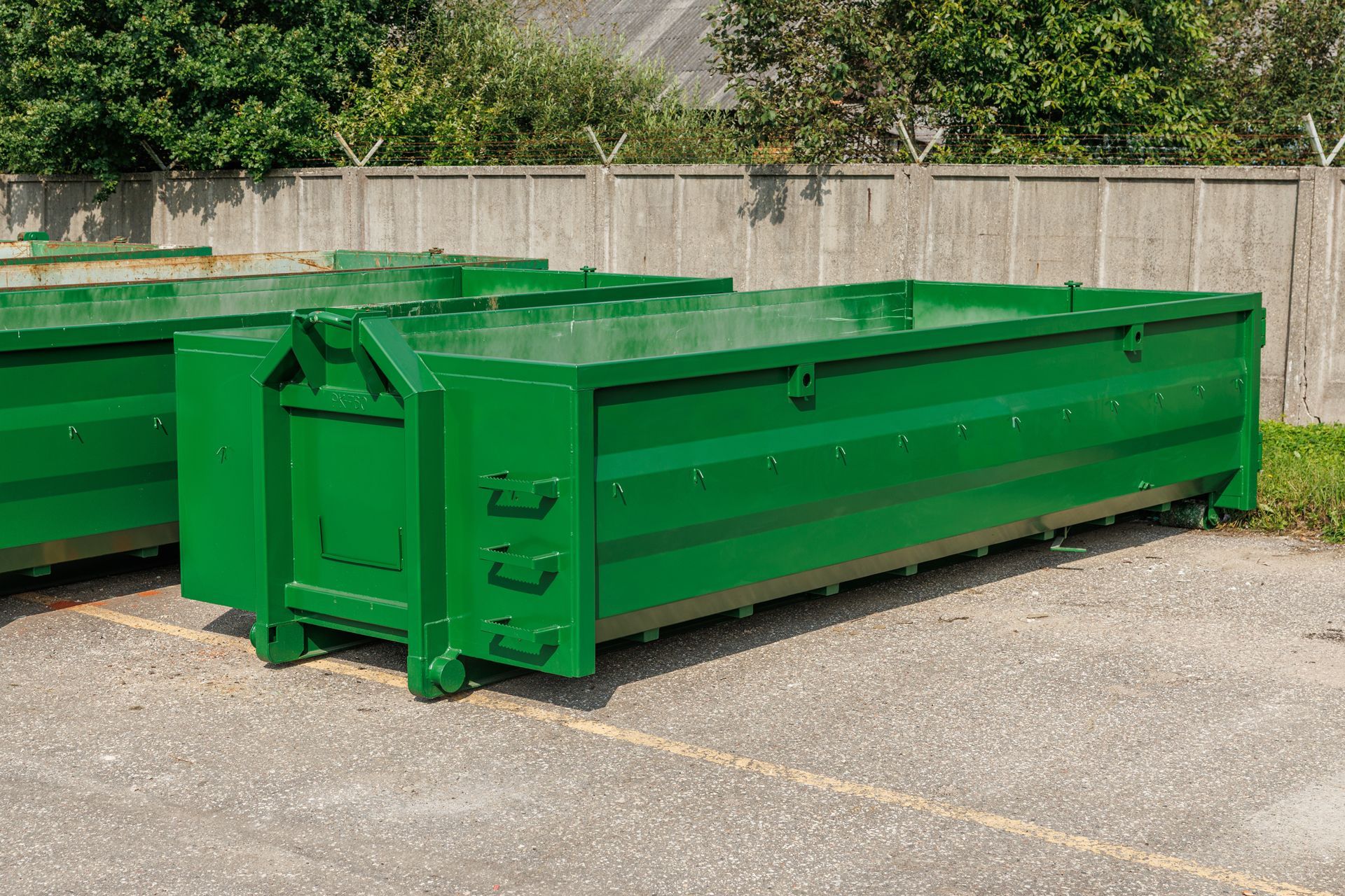 Two empty green metal dumpsters parked on a paved lot in front of a gray fence and trees.