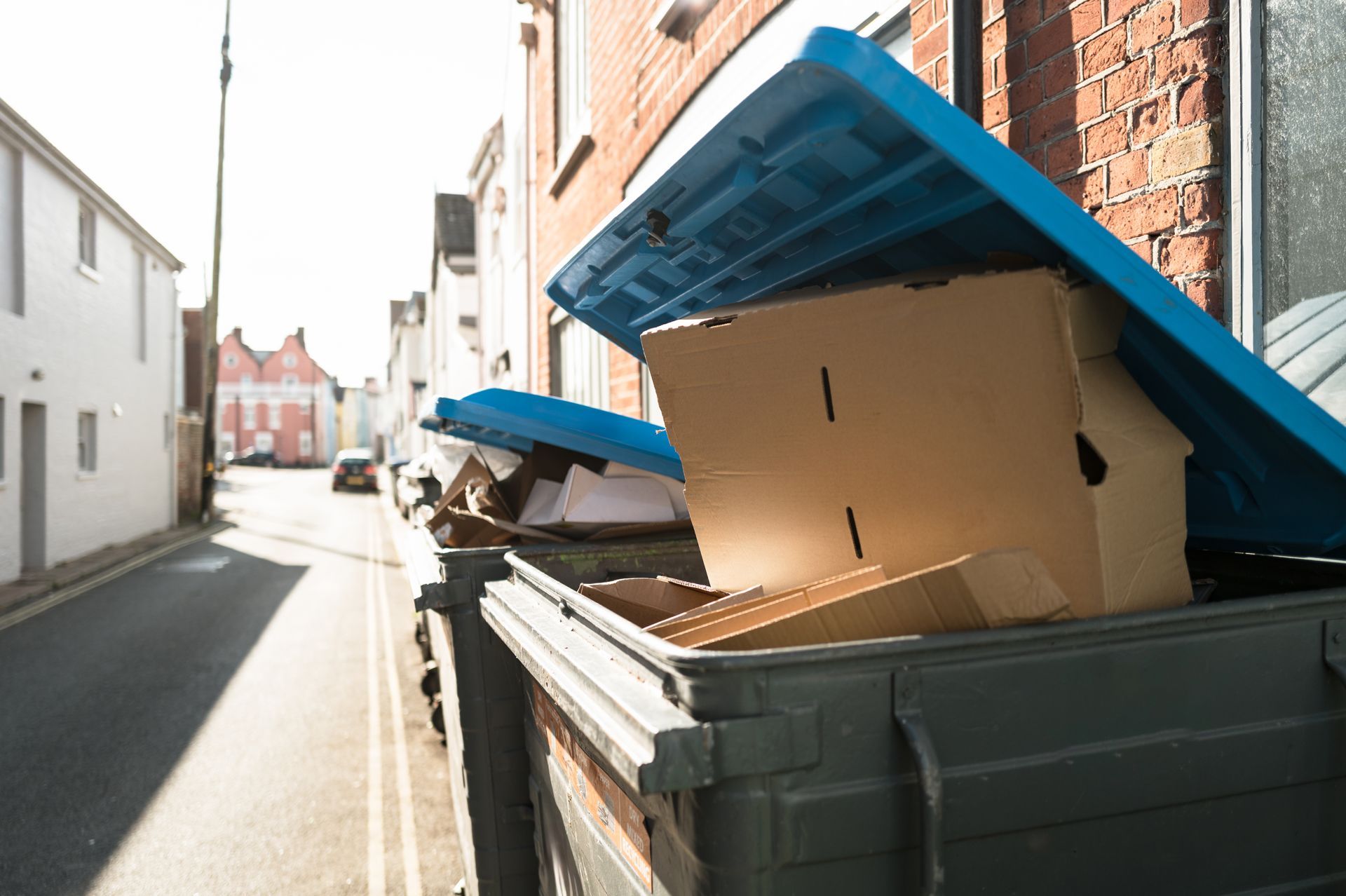 A large, grey waste container with an open blue lid, overflowing with flattened cardboard boxes on a narrow urban street.