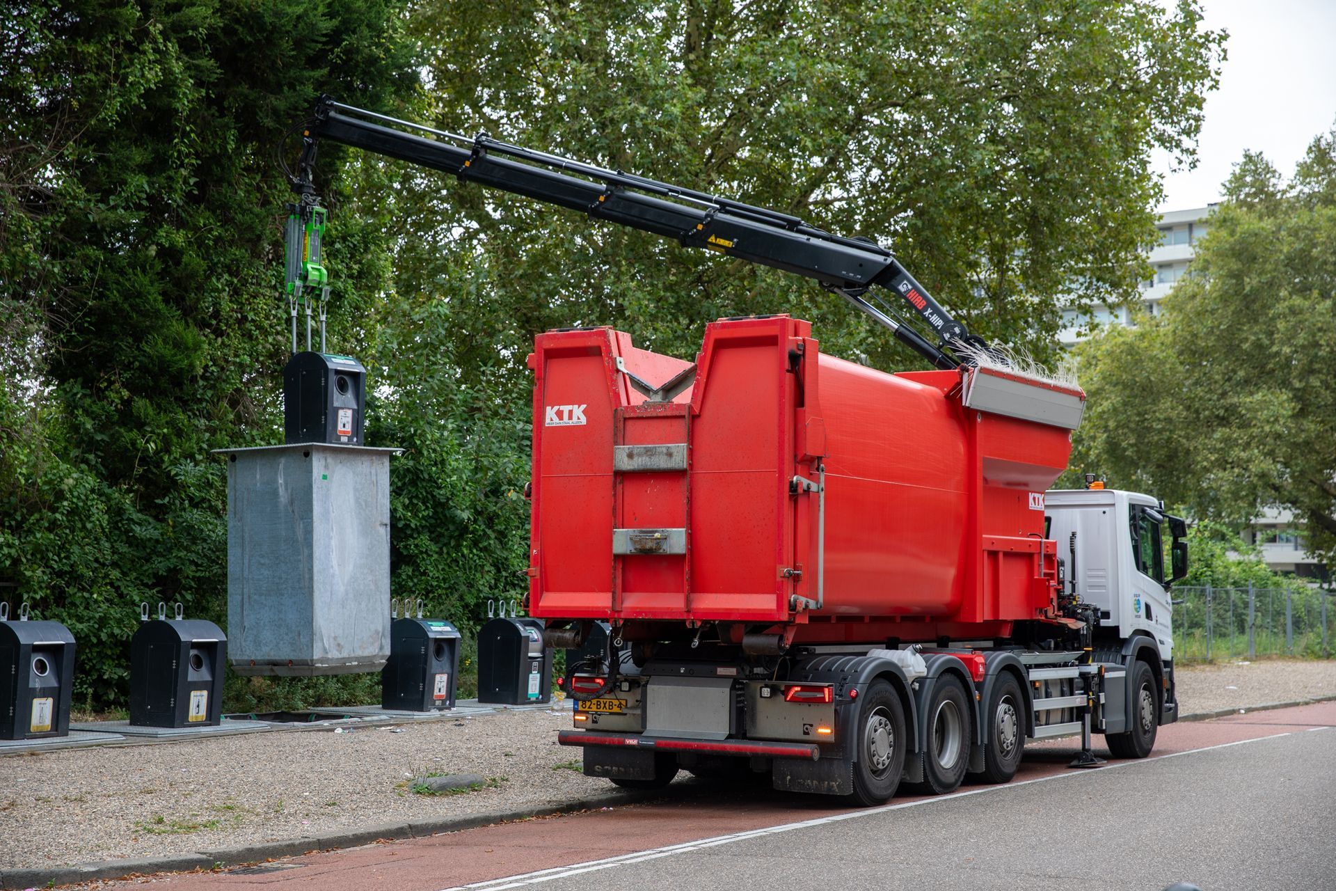 A bright red crane truck lifts a metal container from a recycling station on a roadside.
