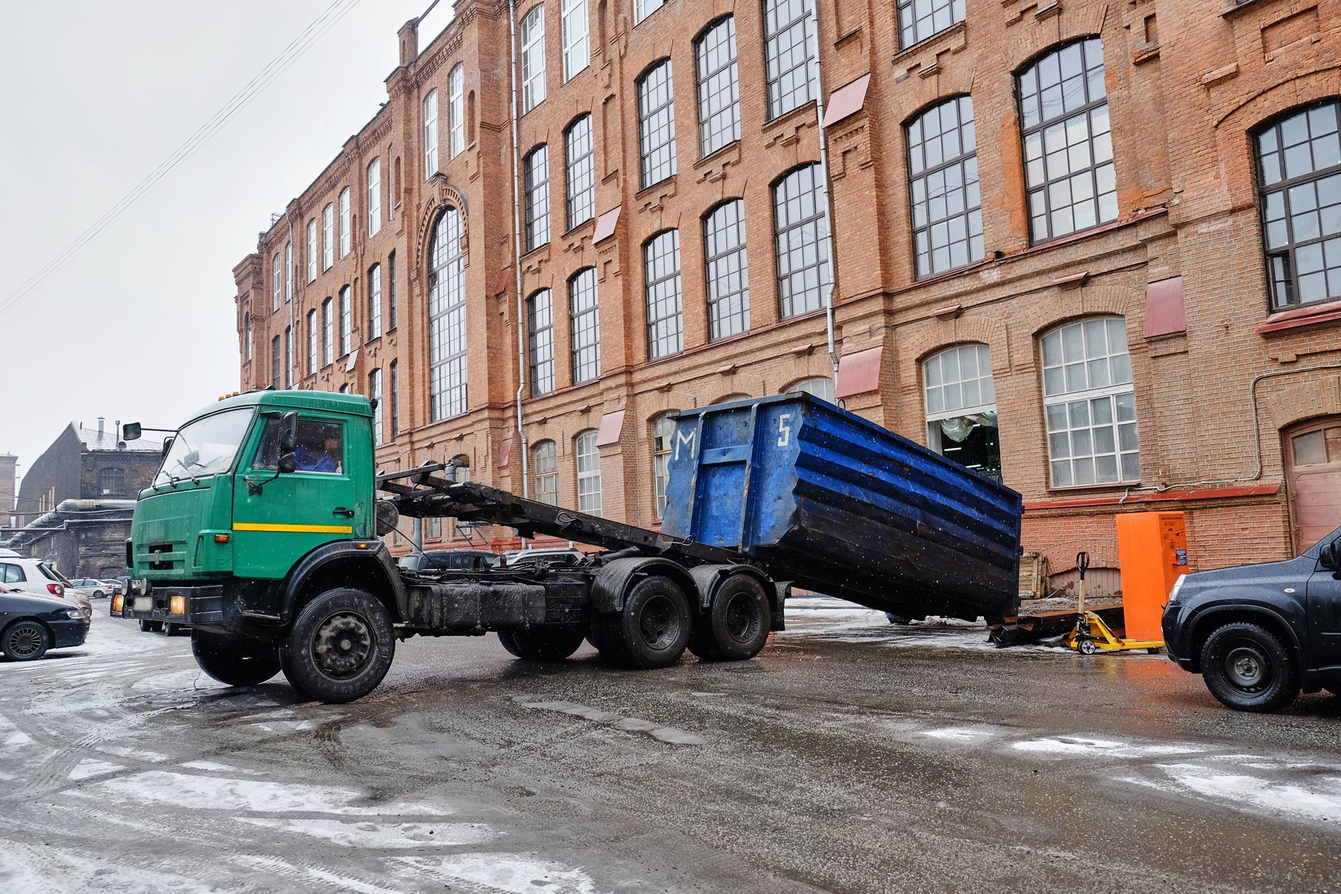 A green roll-off truck positioning a large blue container in front of a red brick industrial building on a snowy day.