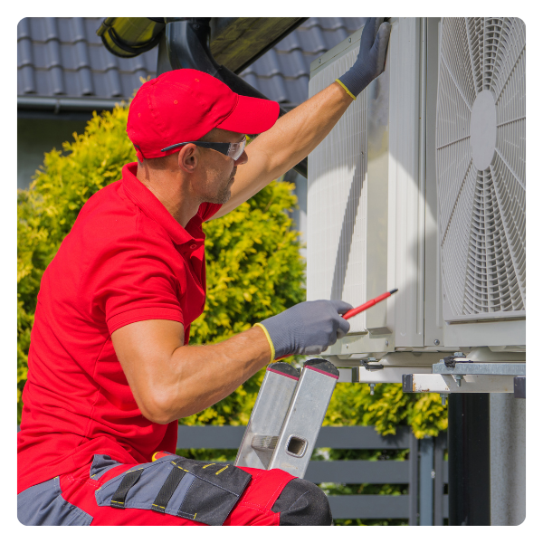 HVAC technician in red shirt and cap on ladder, repairing outdoor unit, holding screwdriver.