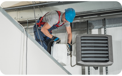 Construction worker in harness installing HVAC unit on a building wall.