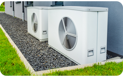 Two white heat pump units sit on gravel next to a building with green grass border.