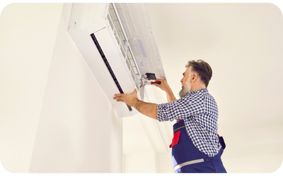 A technician in overalls repairs a wall-mounted air conditioning unit.