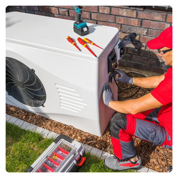 HVAC technician in red shirt and gray pants repairing an outdoor heat pump unit; tools nearby.