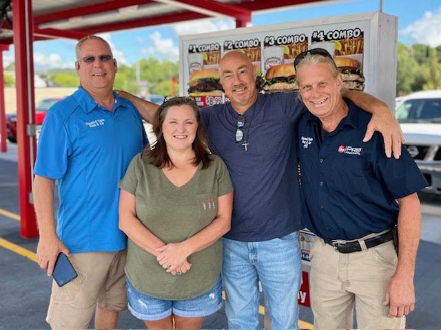 Four people pose by a fast food menu. Two men have their arms around two others. Bright sunlight.