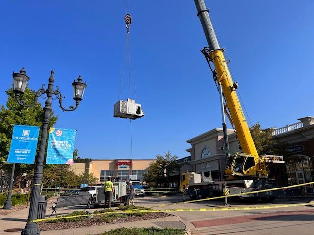 A crane lifting a large, square, stone structure in front of a shopping center on a sunny day.