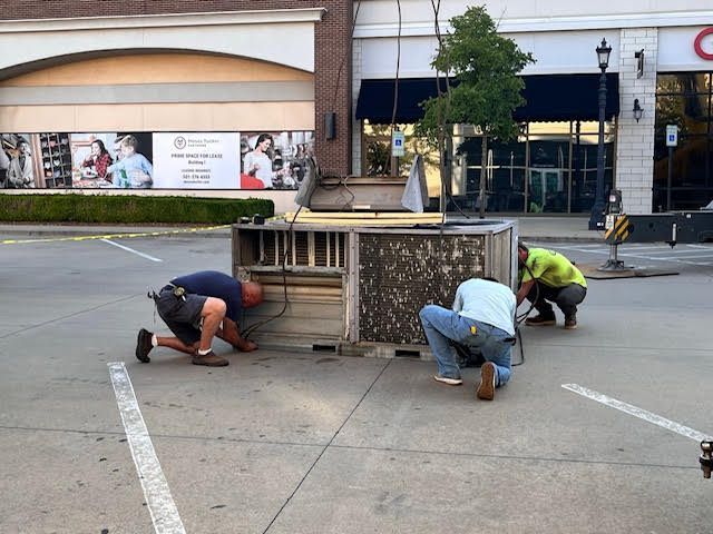 Three workers repairing a wooden structure around a tree in a parking lot. Buildings in background.