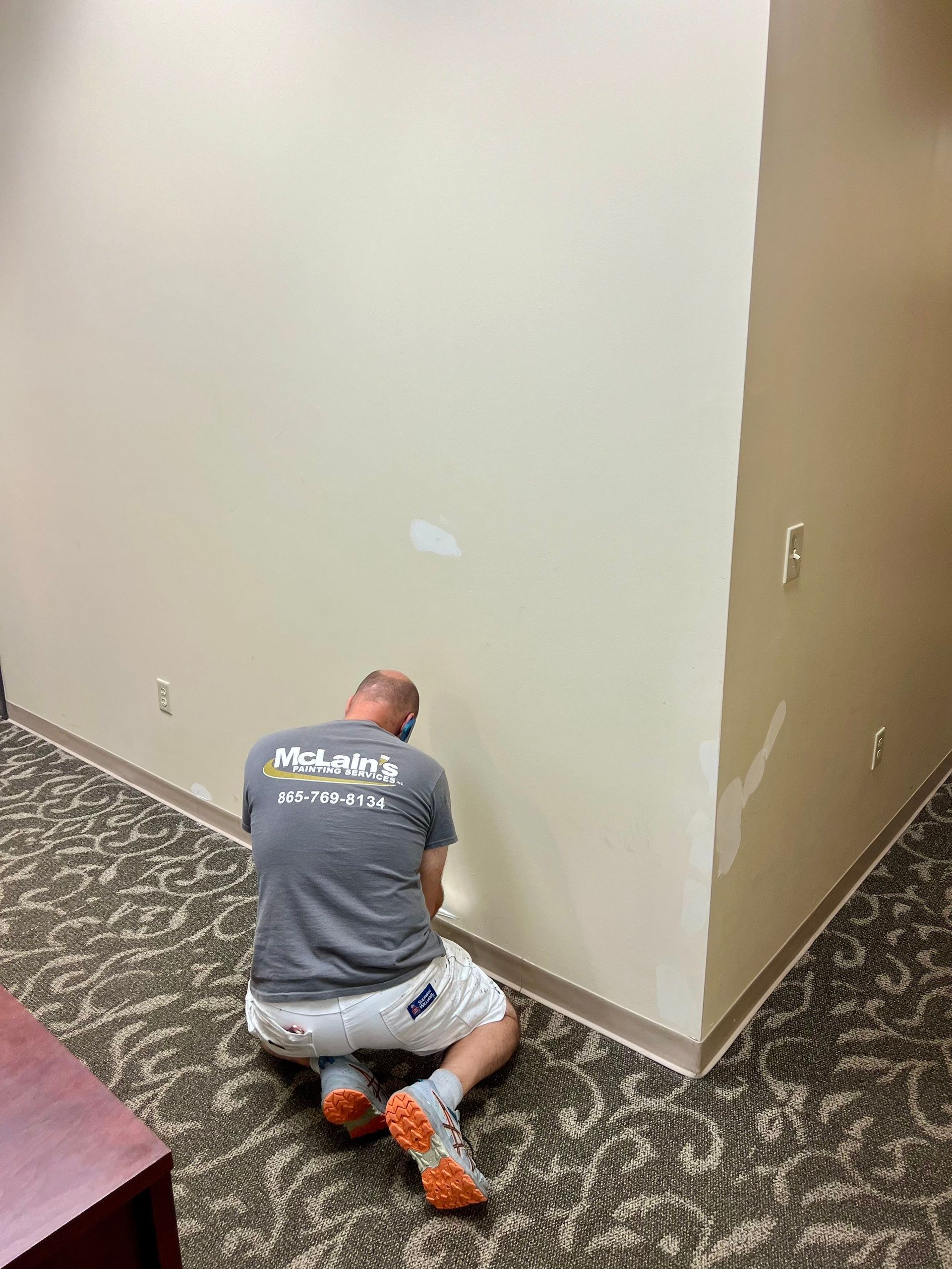 A man is kneeling down in a room painting a wall.