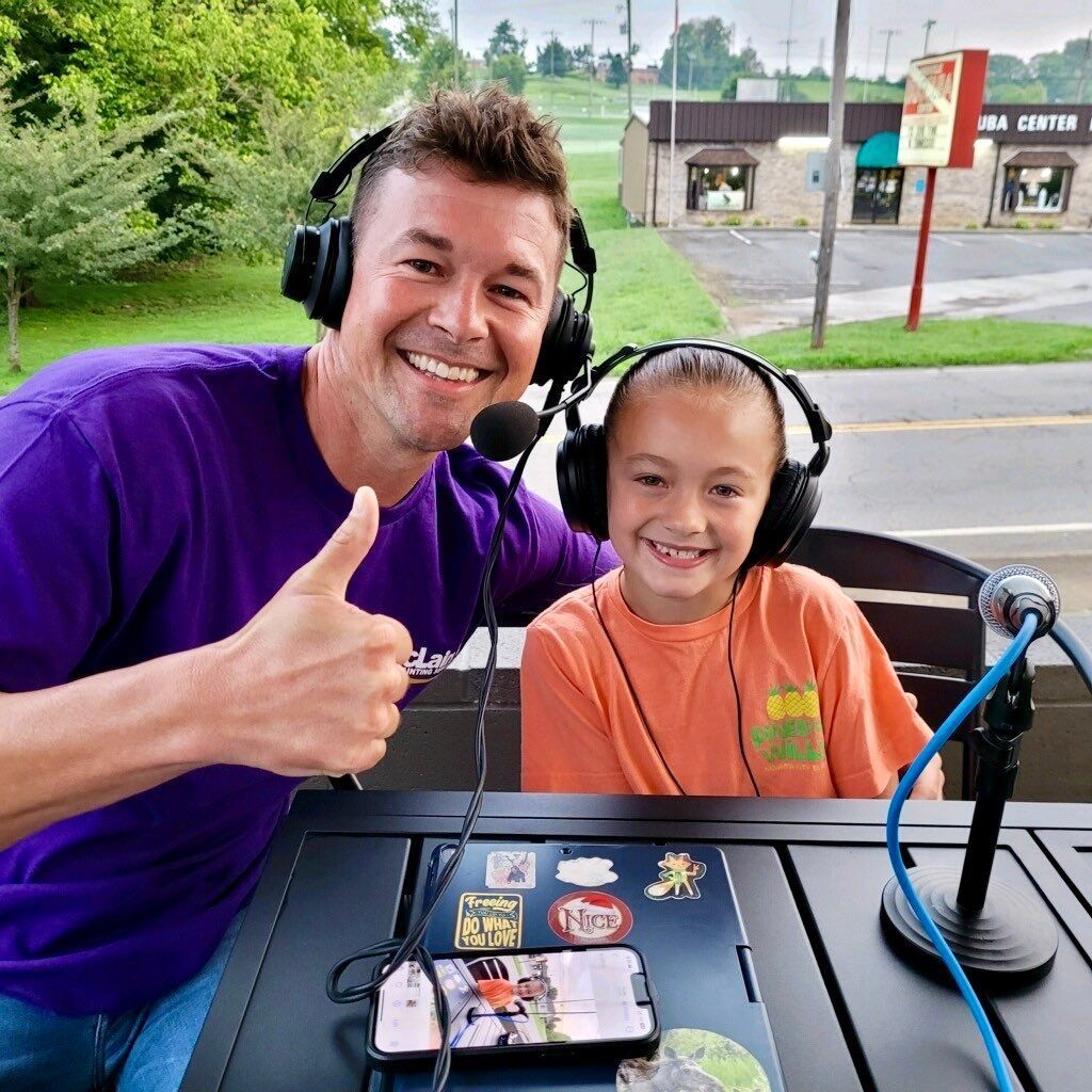 A man and a little girl are sitting at a table wearing headphones and giving a thumbs up.